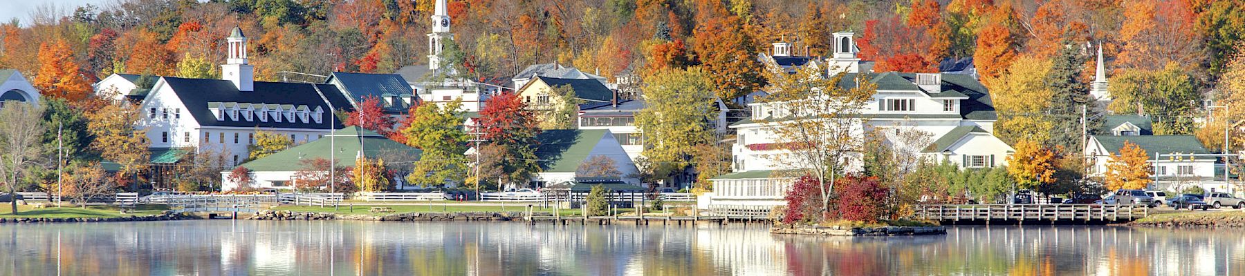 Colorful autumn village by calm water, reflections mirror the orange and yellow trees and white buildings under a clear sky.