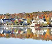 Colorful autumn village by calm water, reflections mirror the orange and yellow trees and white buildings under a clear sky.