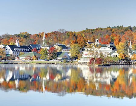 Colorful autumn village by calm water, reflections mirror the orange and yellow trees and white buildings under a clear sky.