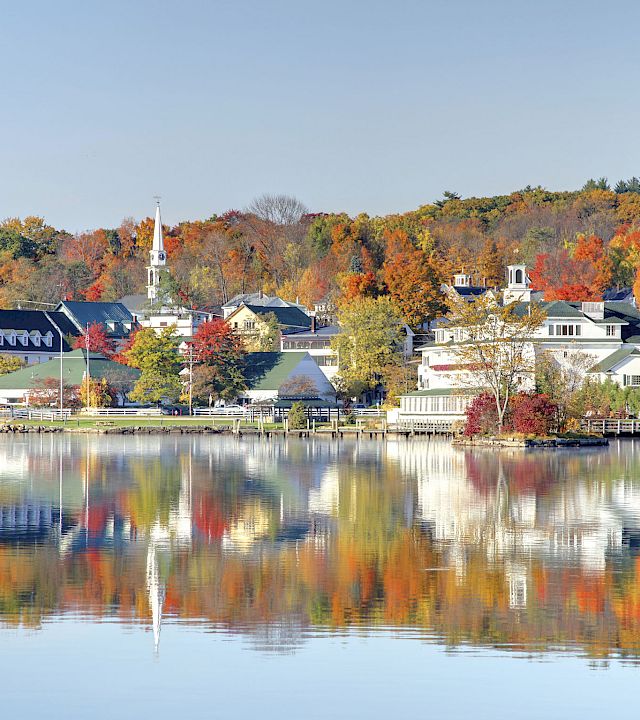 Colorful autumn village by calm water, reflections mirror the orange and yellow trees and white buildings under a clear sky.