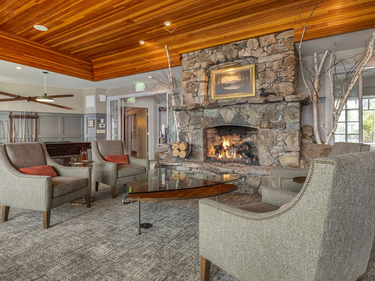 A cozy lounge area in the lobby of Church Landing at Mill Falls with a stone fireplace, wooden ceiling, and multiple gray chairs arranged around a coffee table inside a spacious, well-lit room.