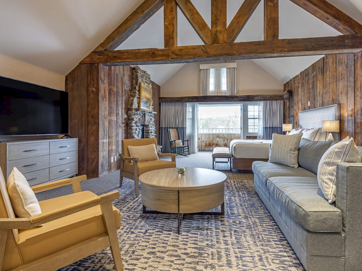 Rustic loft living room with exposed beams, sofa, chairs, TV, stone fireplace, and a round coffee table by large windows.