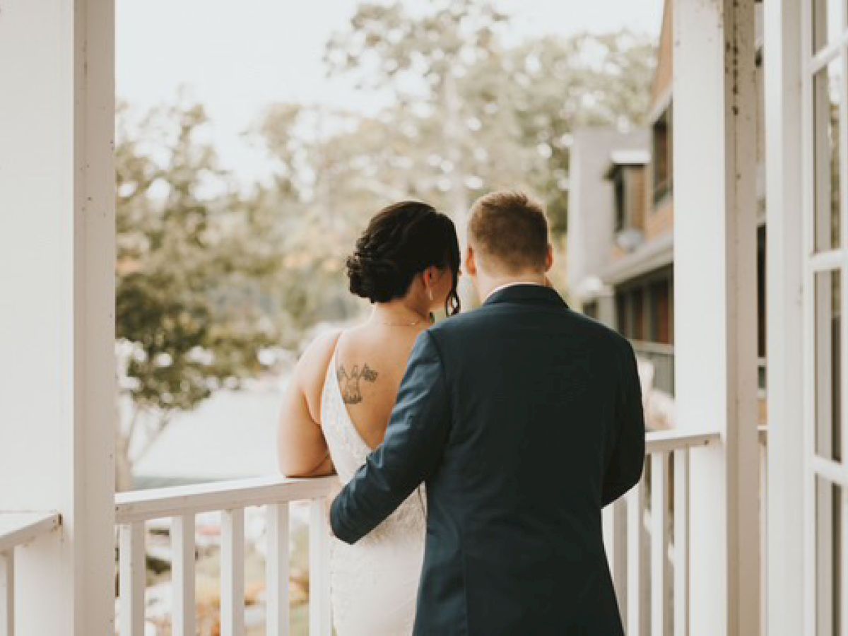 A couple, dressed formally, stands on a balcony overlooking a scenic view, with trees and buildings in the background.