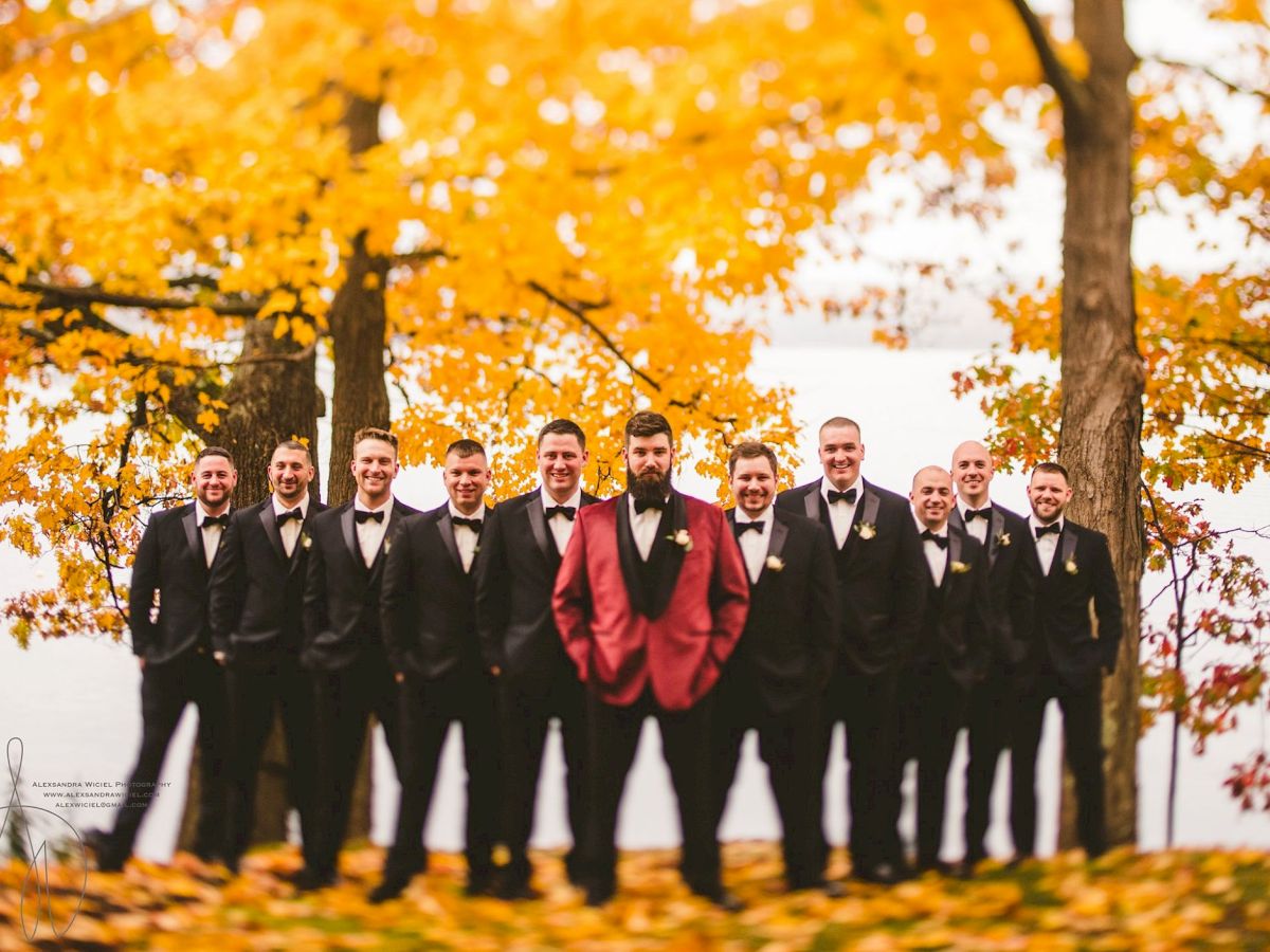 A group of groomsmen in black tuxedos pose outdoors in fall leaves, with one man in a red jacket, celebrating a wedding day.