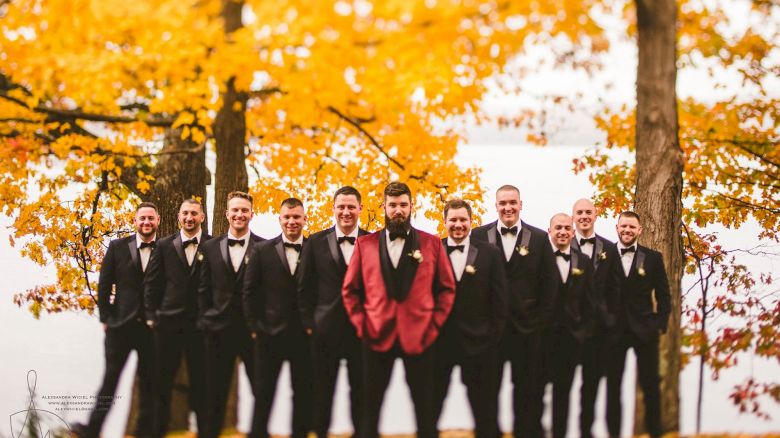 A group of groomsmen in black tuxedos pose outdoors in fall leaves, with one man in a red jacket, celebrating a wedding day.