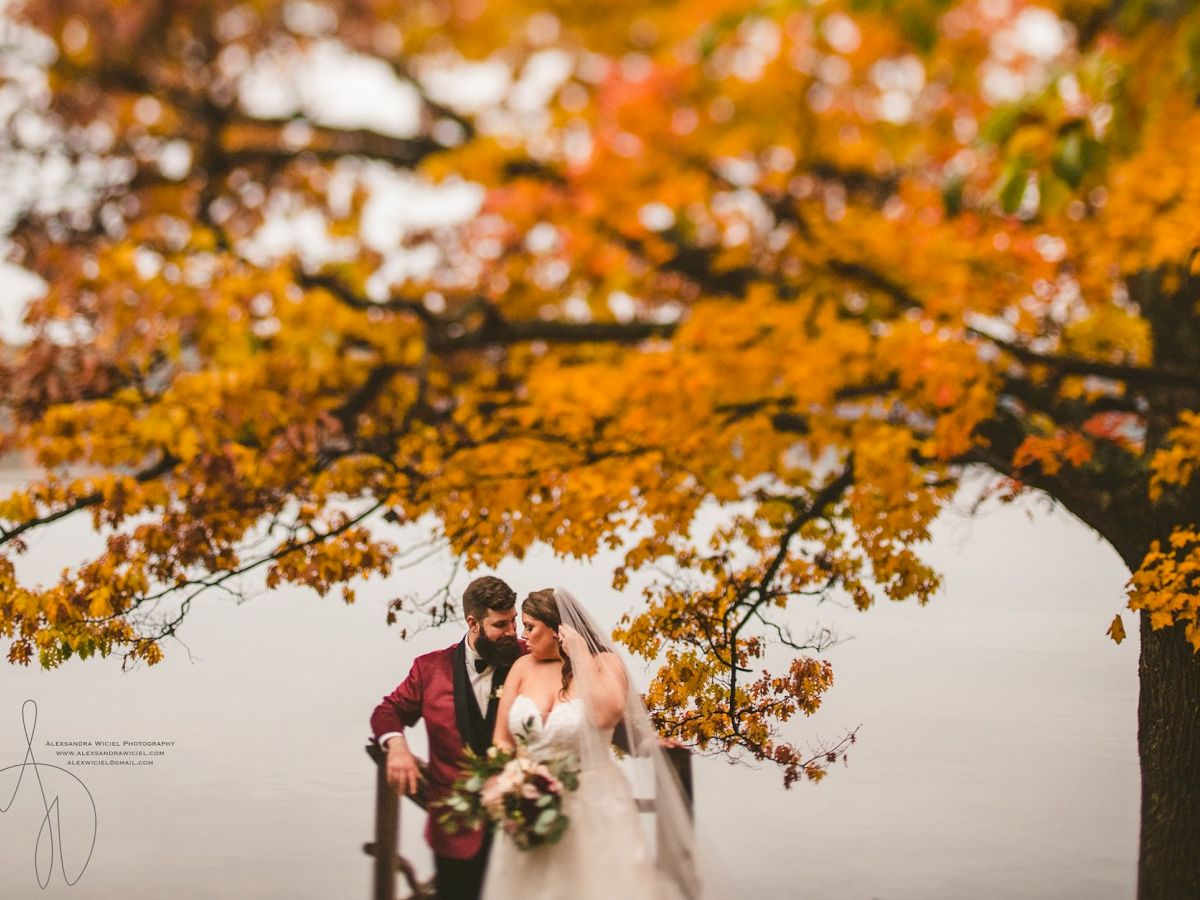 A bride and groom stand together outside in autumn trees, framed by bright orange and yellow leaves, smiling softly.
