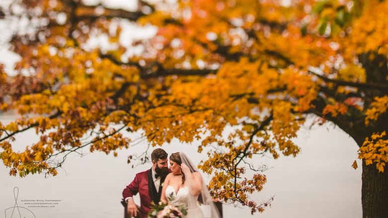 A bride and groom stand together outside in autumn trees, framed by bright orange and yellow leaves, smiling softly.