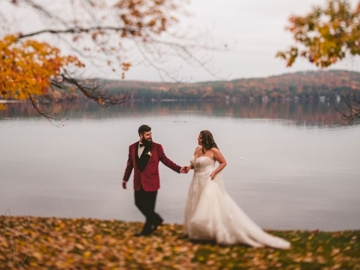 A bride and groom in formalwear stroll by a calm lake in autumn, holding hands with trees and hills behind them.