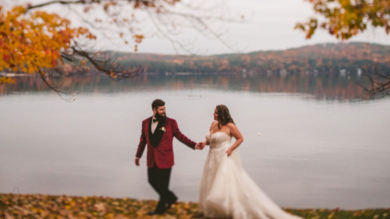 A bride and groom in formalwear stroll by a calm lake in autumn, holding hands with trees and hills behind them.