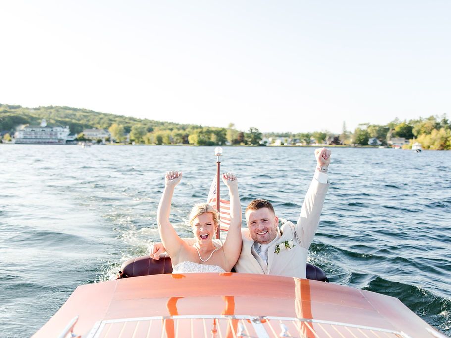 Two people cheer on a vintage wooden ChrisCraft boat as it sails on a calm lake, with a green shoreline and clear sky in the background.