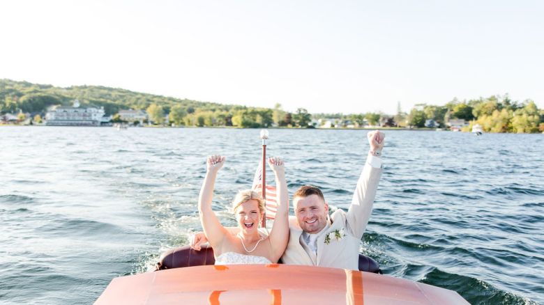 Two people cheer on a vintage wooden ChrisCraft boat as it sails on a calm lake, with a green shoreline and clear sky in the background.
