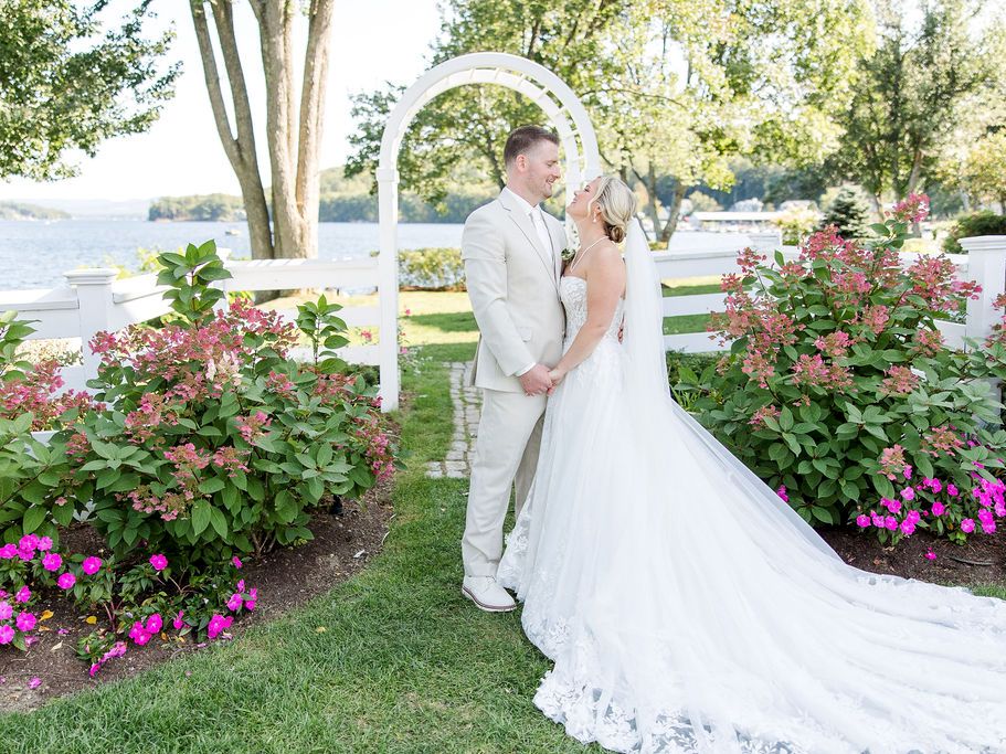 A newlywed couple in wedding attire share a kiss under a white archway with Lake Winnipesaukee in background, surrounded by colorful flowers and greenery on a sunny day.