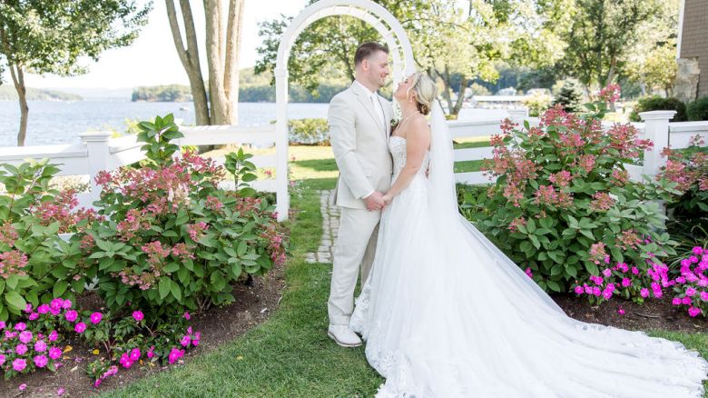 A newlywed couple in wedding attire share a kiss under a white archway with Lake Winnipesaukee in background, surrounded by colorful flowers and greenery on a sunny day.