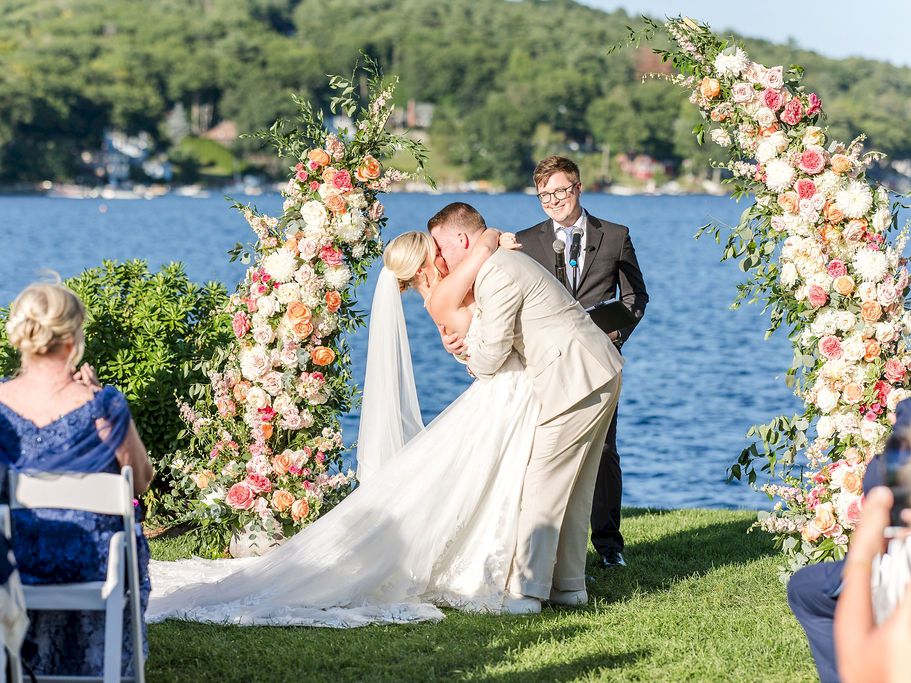 A couple kissing at their outdoor wedding under a floral arch by a lake, with guests watching on.