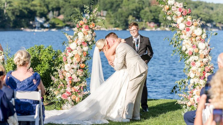 A couple kissing at their outdoor wedding under a floral arch by a lake, with guests watching on.