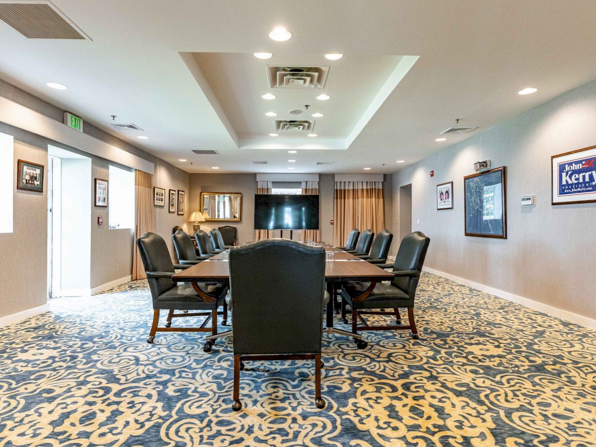 A conference room with a long table, black chairs, framed posters on the wall, ornate blue carpet, and ceiling lights.