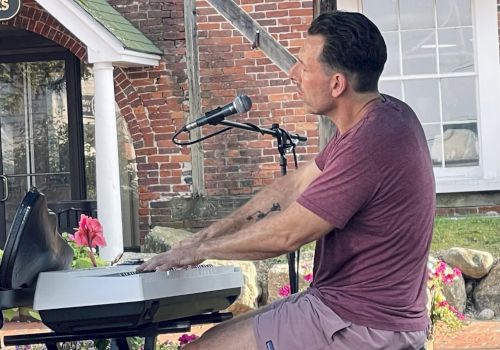 A man plays a keyboard and sings into a microphone outdoors on a patio, with a brick wall backdrop and flowers nearby.