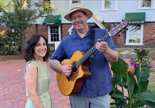 A smiling guitarist in a hat holds a yellow acoustic guitar with a woman in a green dress beside him outdoors near colorful flowers.