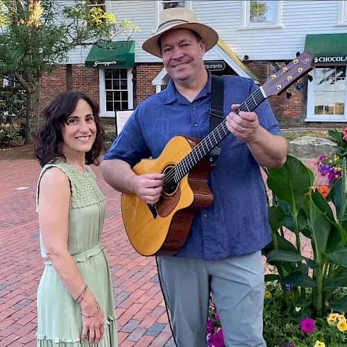 A smiling guitarist in a hat holds a yellow acoustic guitar with a woman in a green dress beside him outdoors near colorful flowers.