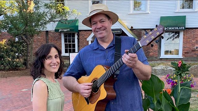 A smiling guitarist in a hat holds a yellow acoustic guitar with a woman in a green dress beside him outdoors near colorful flowers.