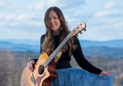 A woman outdoors smiling and playing an acoustic guitar, standing in a scenic mountain landscape.