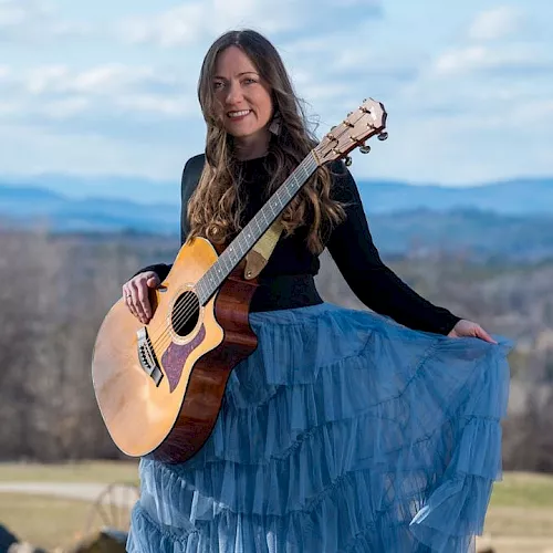 A woman outdoors smiling and playing an acoustic guitar, standing in a scenic mountain landscape.