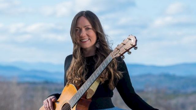 A woman outdoors smiling and playing an acoustic guitar, standing in a scenic mountain landscape.