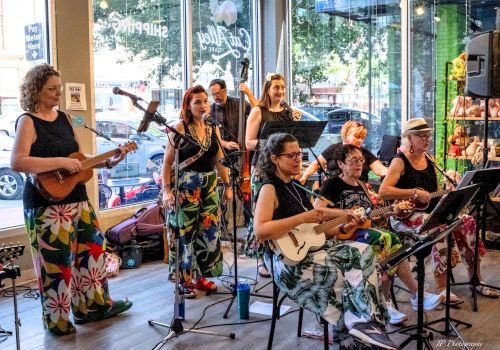 A group of women performs live music indoors, singing into microphones and playing guitars at a casual gathering.