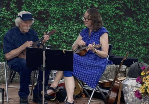 An elderly man plays a ukulele while a woman sings or reads music outdoors, with other string instruments and flowers nearby.