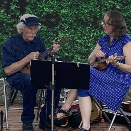 An elderly man plays a ukulele while a woman sings or reads music outdoors, with other string instruments and flowers nearby.