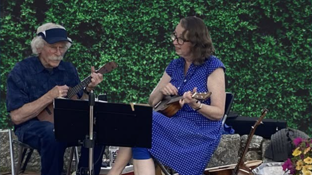 An elderly man plays a ukulele while a woman sings or reads music outdoors, with other string instruments and flowers nearby.