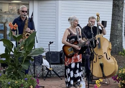 An outdoor band is performing on a porch with musicians playing guitar, bass, and vocals beside flowers and speakers.