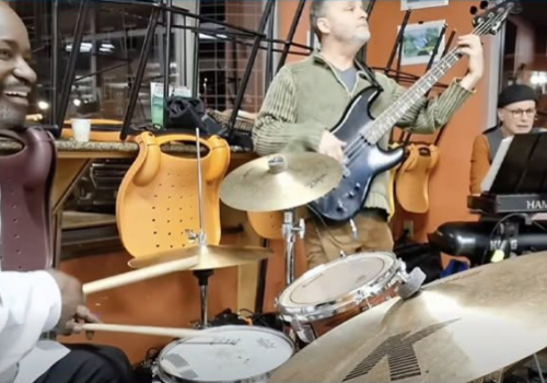 A group of musicians rehearses in a studio: a drummer, guitarist, keyboardist, and others with instruments under warm lights.