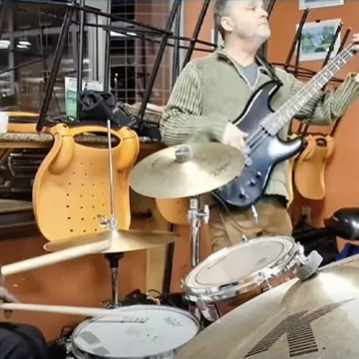 A group of musicians rehearses in a studio: a drummer, guitarist, keyboardist, and others with instruments under warm lights.