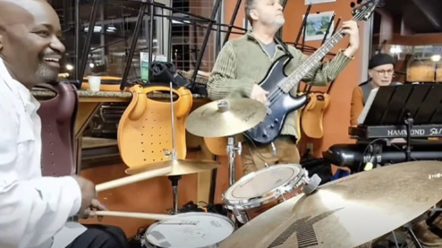 A group of musicians rehearses in a studio: a drummer, guitarist, keyboardist, and others with instruments under warm lights.