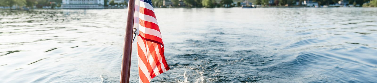 View from a boat with an American flag waving at the bow as it cruises over rippling water toward shore.