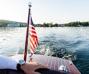 View from a boat with an American flag waving at the bow as it cruises over rippling water toward shore.