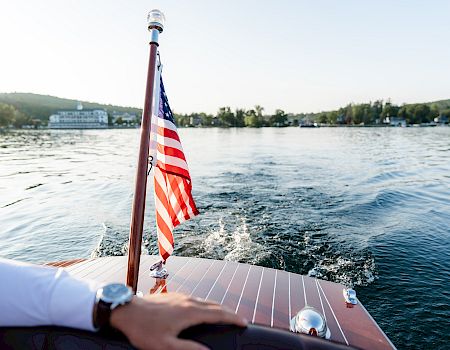 View from a boat with an American flag waving at the bow as it cruises over rippling water toward shore.