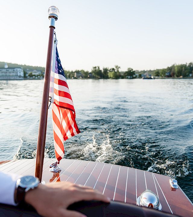 View from a boat with an American flag waving at the bow as it cruises over rippling water toward shore.