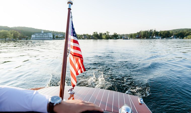View from a boat with an American flag waving at the bow as it cruises over rippling water toward shore.