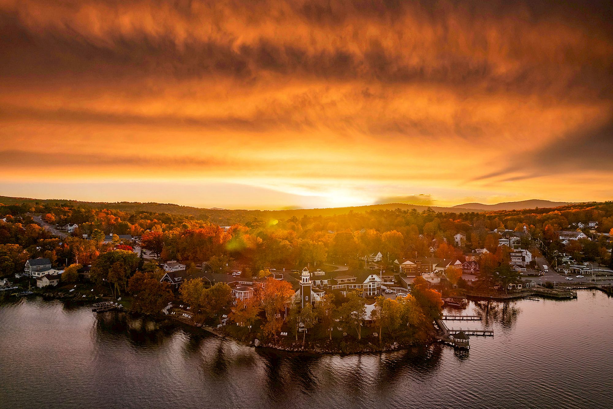 An aerial view of a lakeside village at sunset with a vibrant, fiery sky and scattered houses along the shoreline surrounded by autumn foliage.