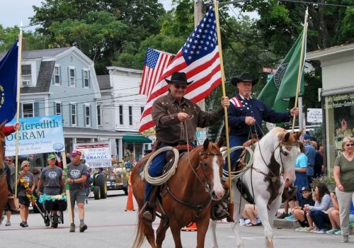 In a town parade, two horse riders wave US flags while spectators watch along Main Street.