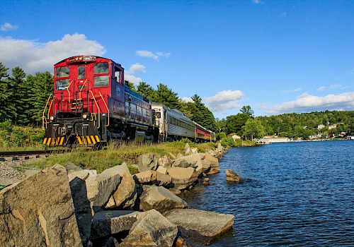 A red locomotive pulls a long passenger train along a scenic lakeshore with trees, rocks, and a bright blue sky.