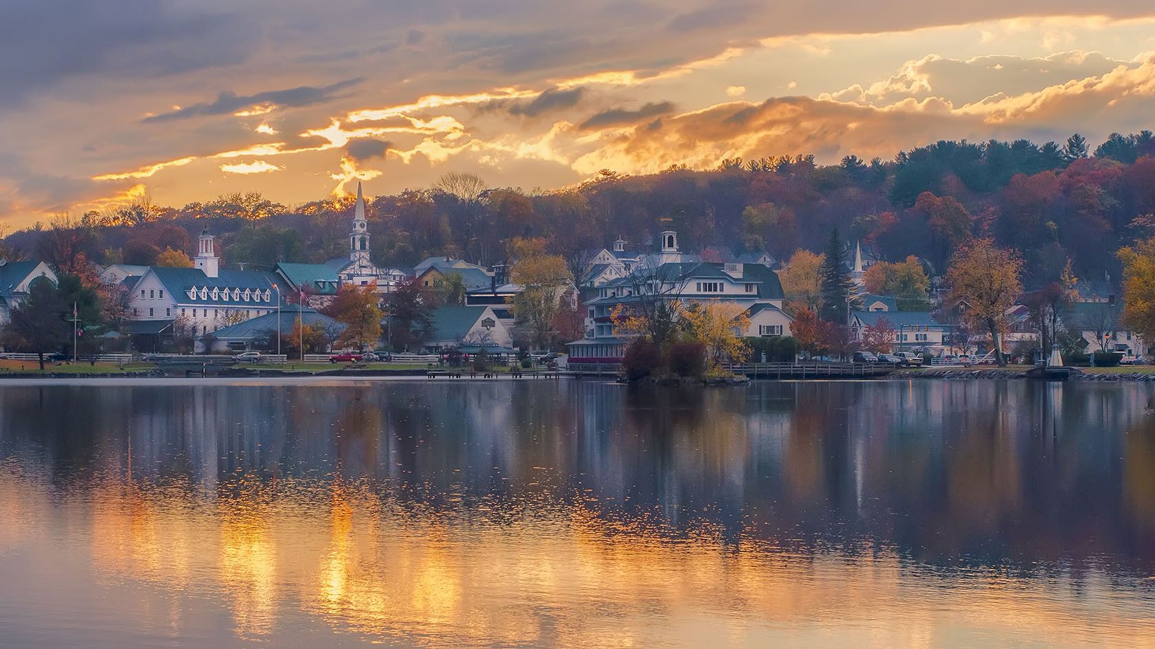 A tranquil lakeside town at sunset, with orange skies, calm water, and colorful autumn trees reflecting off the reflective surface.