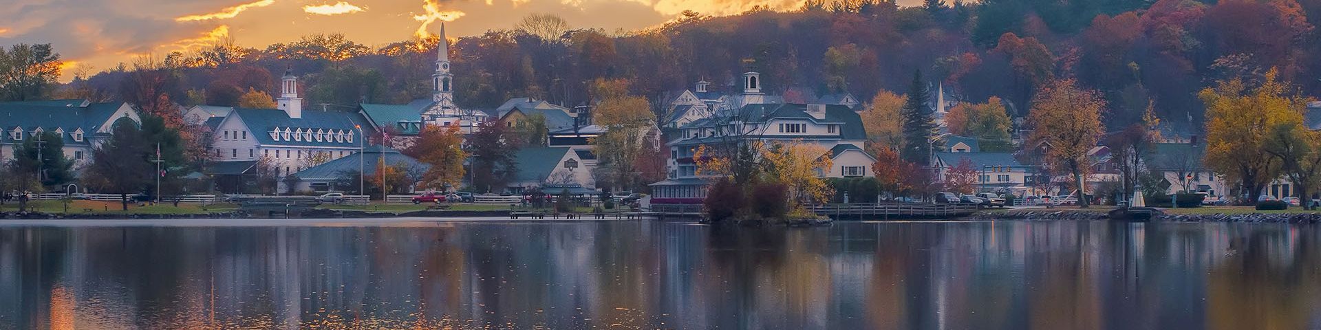 A tranquil lakeside town at sunset, with orange skies, calm water, and colorful autumn trees reflecting off the reflective surface.