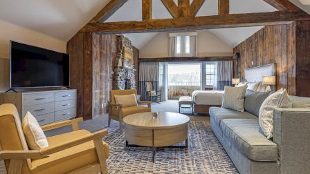A cozy rustic living room with wooden beams, planked walls, a blue-gray sofa, matching chairs, a round coffee table, and a flat-screen TV, bright and inviting.