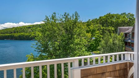 A sunny lakeside balcony with green trees, blue water, and a small table with orange drinks; railing frames the view.