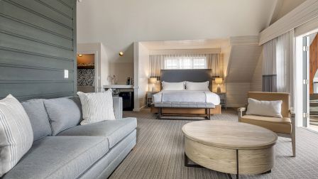 A bright, stylish hotel suite with a gray sofa, round coffee table, and a plush king bed framed by soft lighting and neutral decor, plus a chair and sliding doors to a sunny patio.