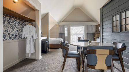 A stylish hotel room with a sitting area, dining table, open wardrobe, a bed in the back, and a balcony overlook, all in neutral tones.