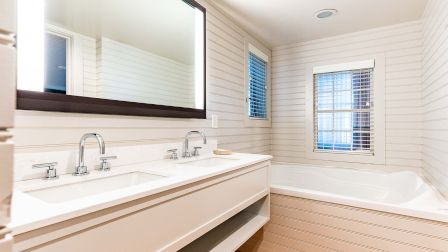 A bright, modern bathroom with a double sink vanity, large wall mirror, white tiled walls, a corner bathtub, and a small window with blinds.